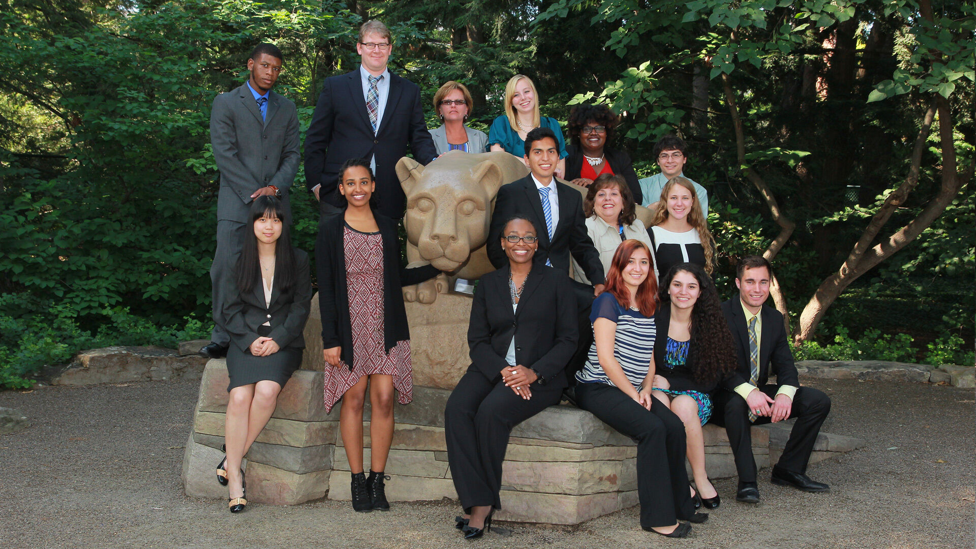 Group of fifteen graduate students posing on a limestone statue of a mountain lion in nice weather with brilliant green foliage in the background