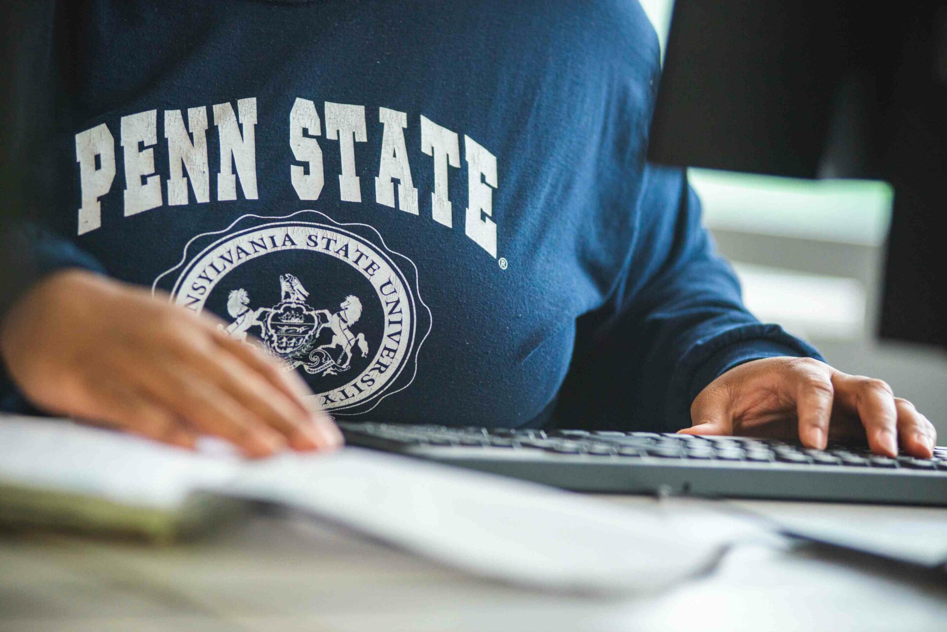 Student wearing a Penn State sweatshirt sitting at a computer with a notebook