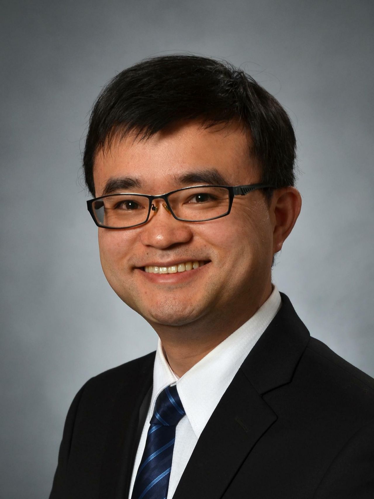 A man with neatly-trimmed brown hair and glasses smiles in a dark jacket, white shirt, and tie.