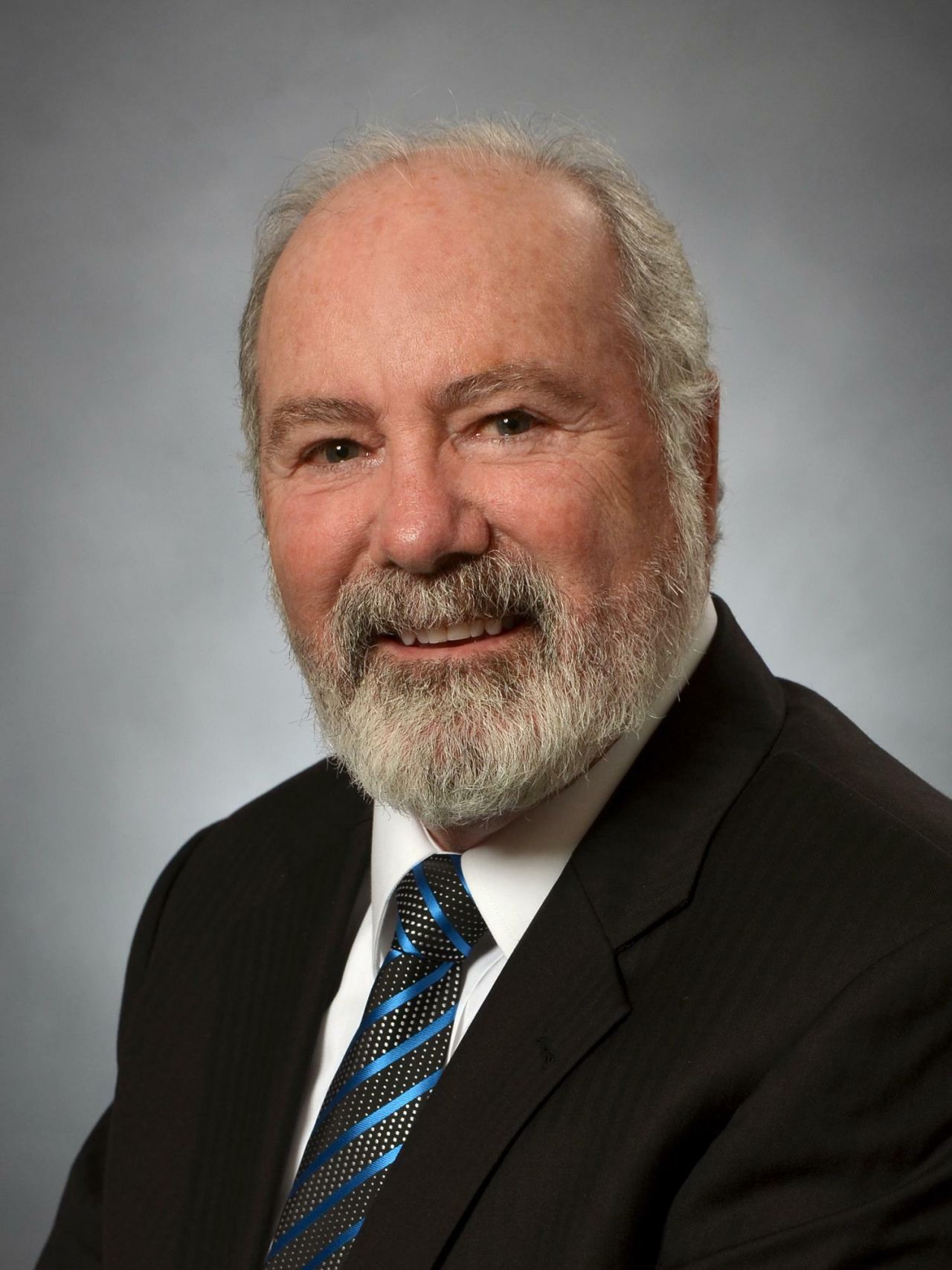 A man with graying beard poses in front of a gray studio backdrop in a dark jacket, white shirt, and tie