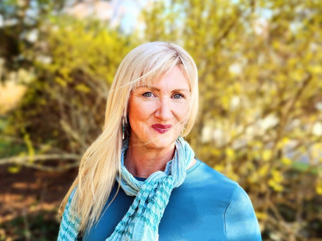 A woman with long blonde hair poses outdoors in front of a softly-focused bright yellow bush. She is wearing a light blue sweater and turquoise scarf.