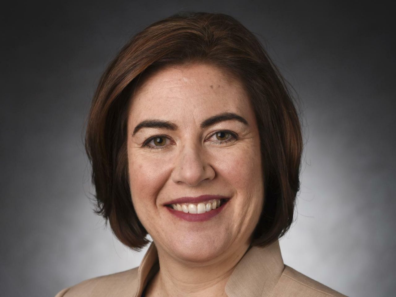 A woman with brown hair in a bob cut smiles broadly in a tan jacket