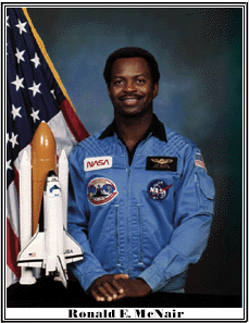 Ronald E. McNair dressed in a NASA suit, standing next to an American flag and a miniature space shuttle model.